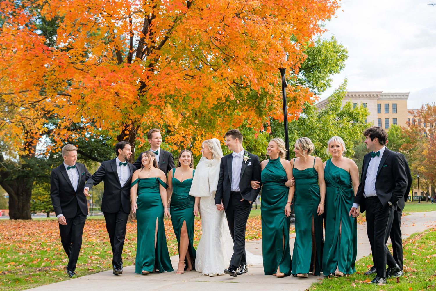 Wedding party poses under vibrant orange autumn trees wearing emerald green and black formal attire.