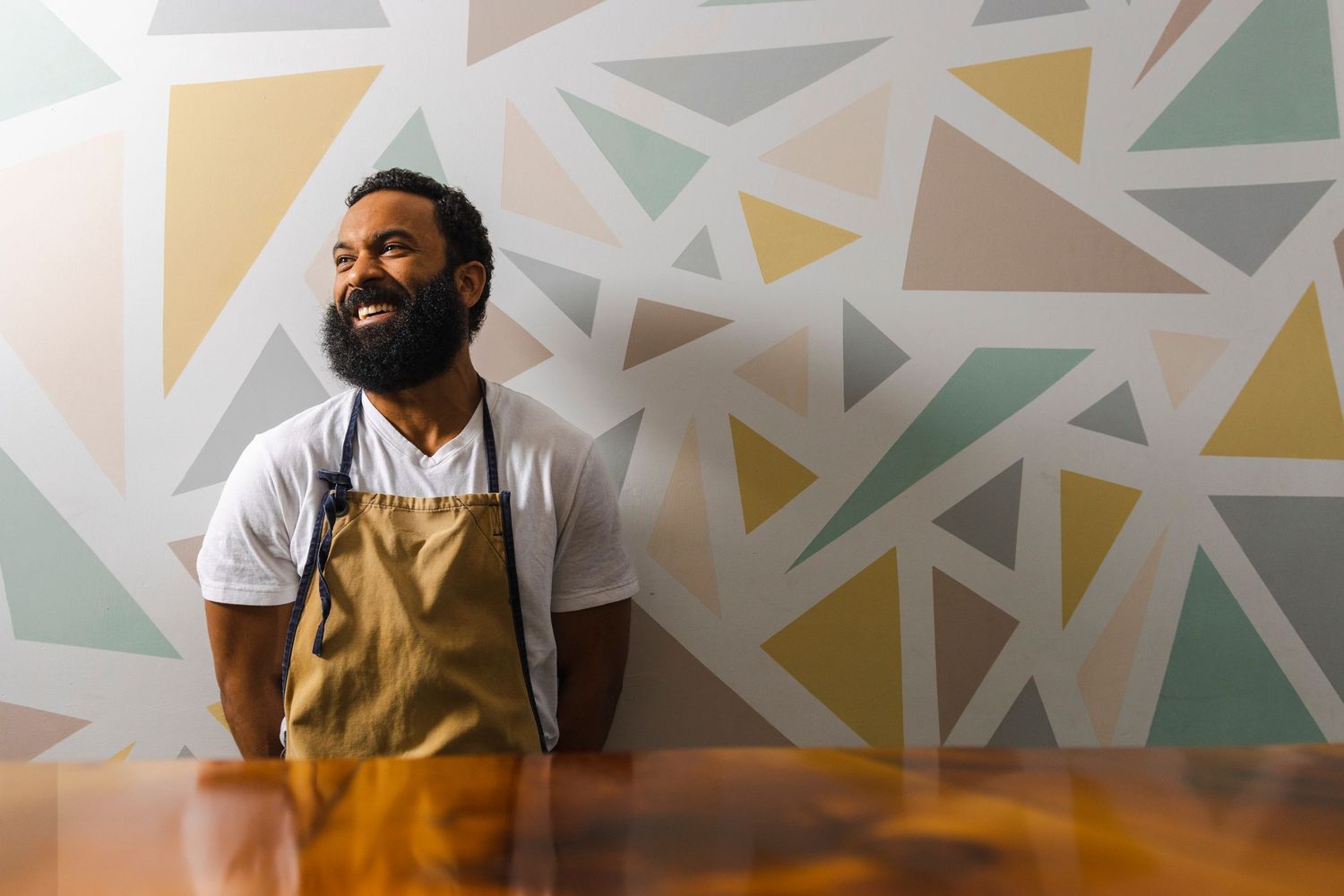 Someone wearing a beige apron and white t-shirt stands against a geometric patterned wall in warm yellow and mint tones.