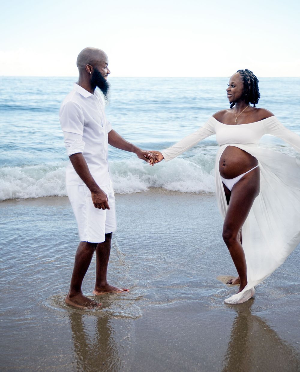 Couple splashing and dancing in ocean waves wearing white attire.