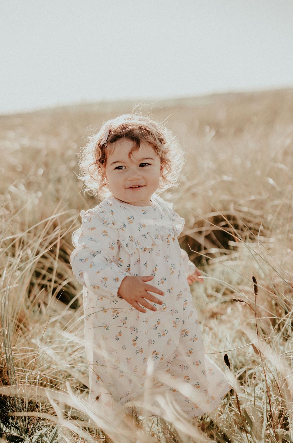 A small child wearing a floral dress plays in a sunlit wheat field during golden hour.