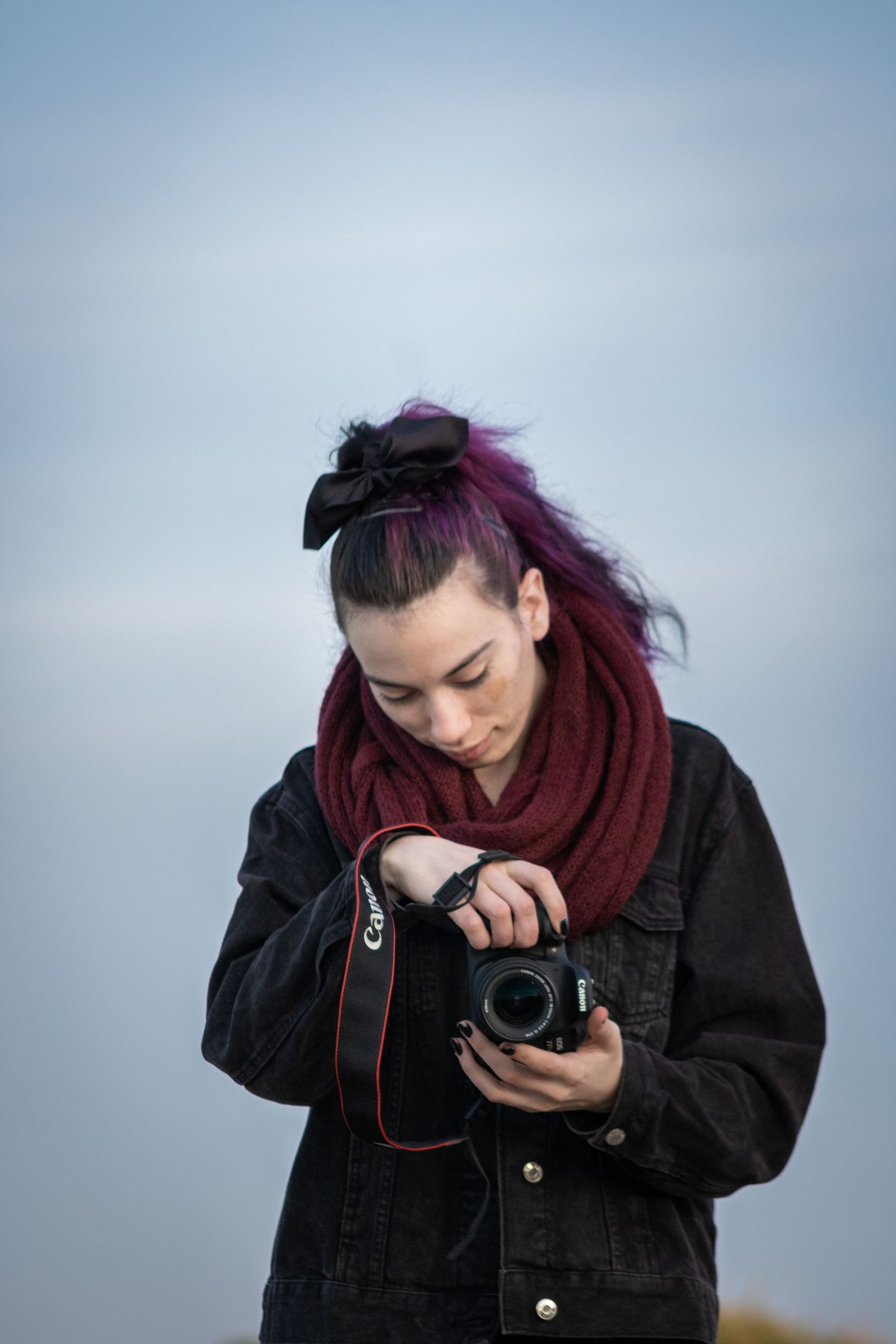 Photographer wearing black jacket and burgundy scarf examines camera settings outdoors.