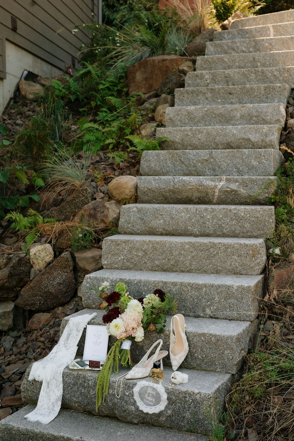 Bridal accessories including heels, bouquet, and lace veil arranged on rustic stone steps surrounded by greenery.