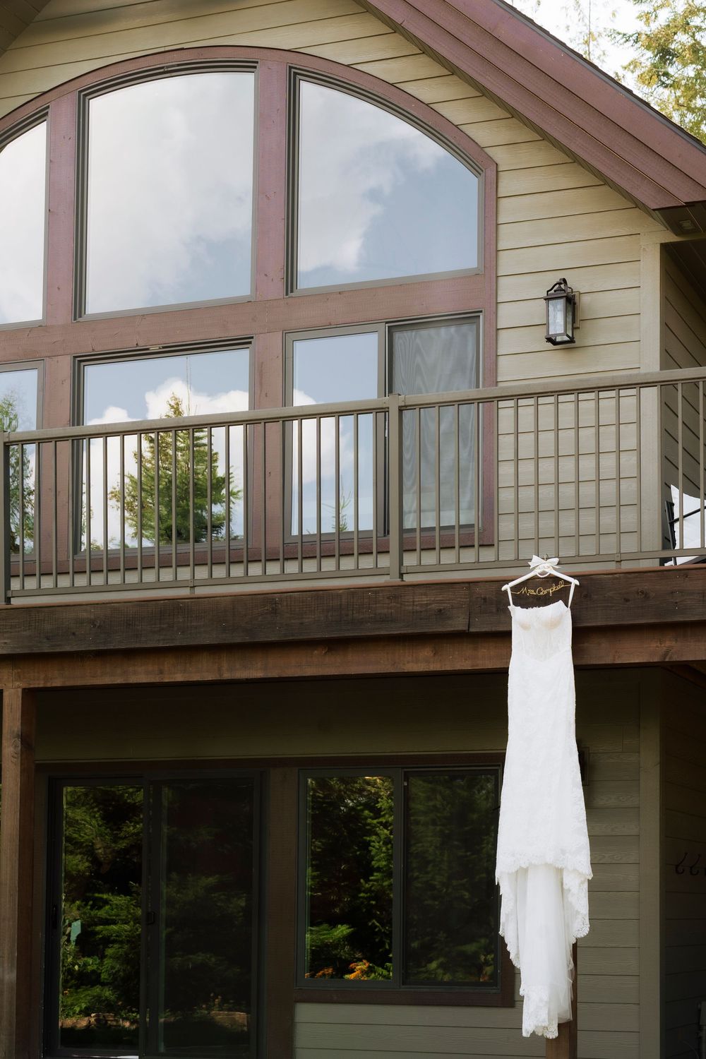 White wedding dress hanging on a hanger outside a rustic cabin with large arched windows and a wooden deck.