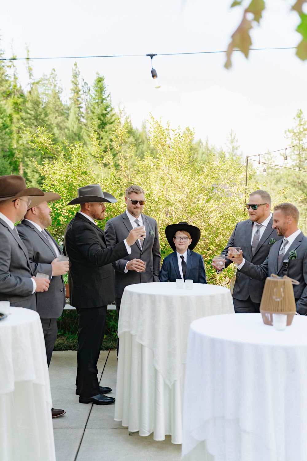Groomsmen in suits and cowboy hats gather outdoors at a wedding cocktail hour with white draped tables.