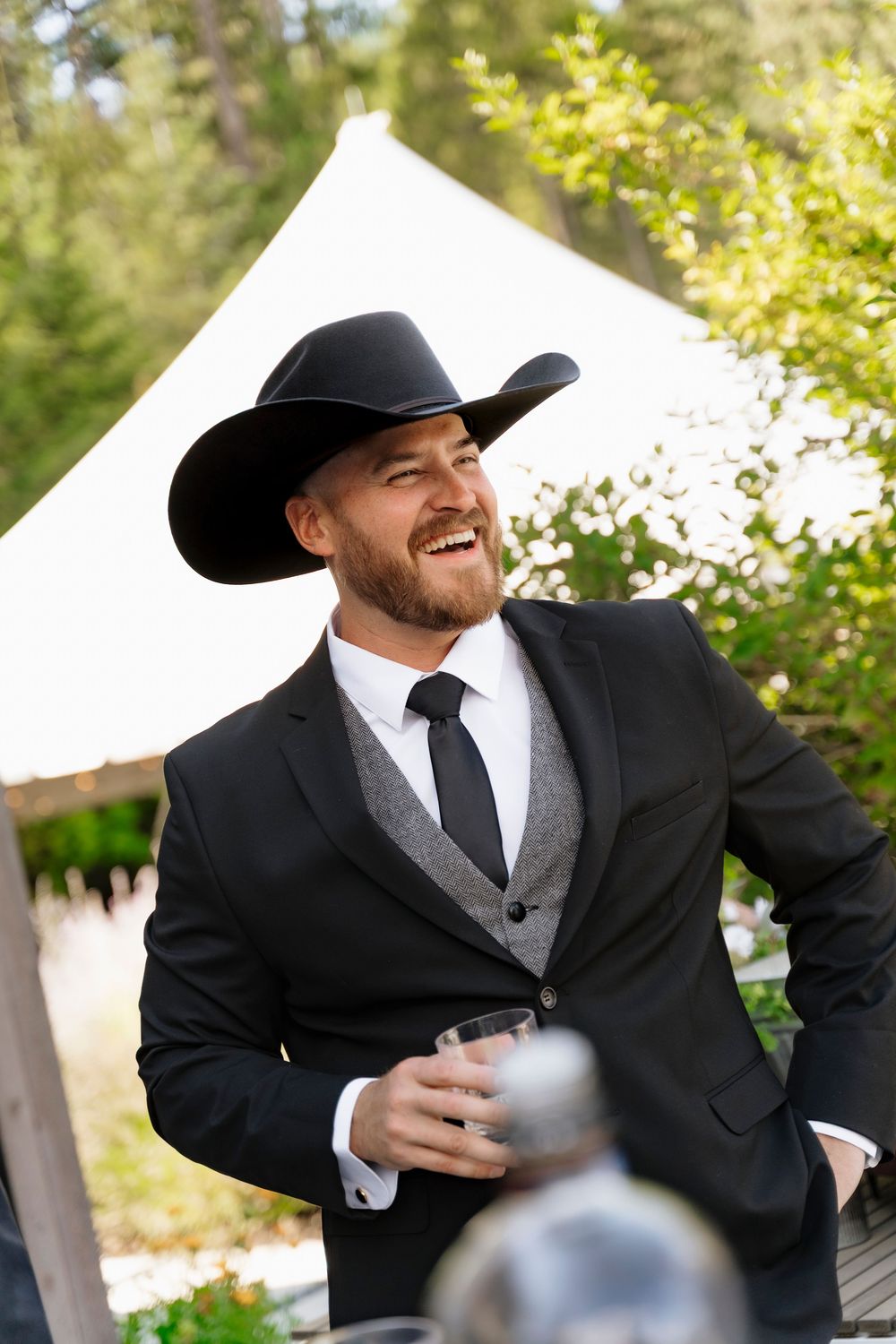 Man in black cowboy hat and suit smiling outdoors at event, holding drink near white tent.