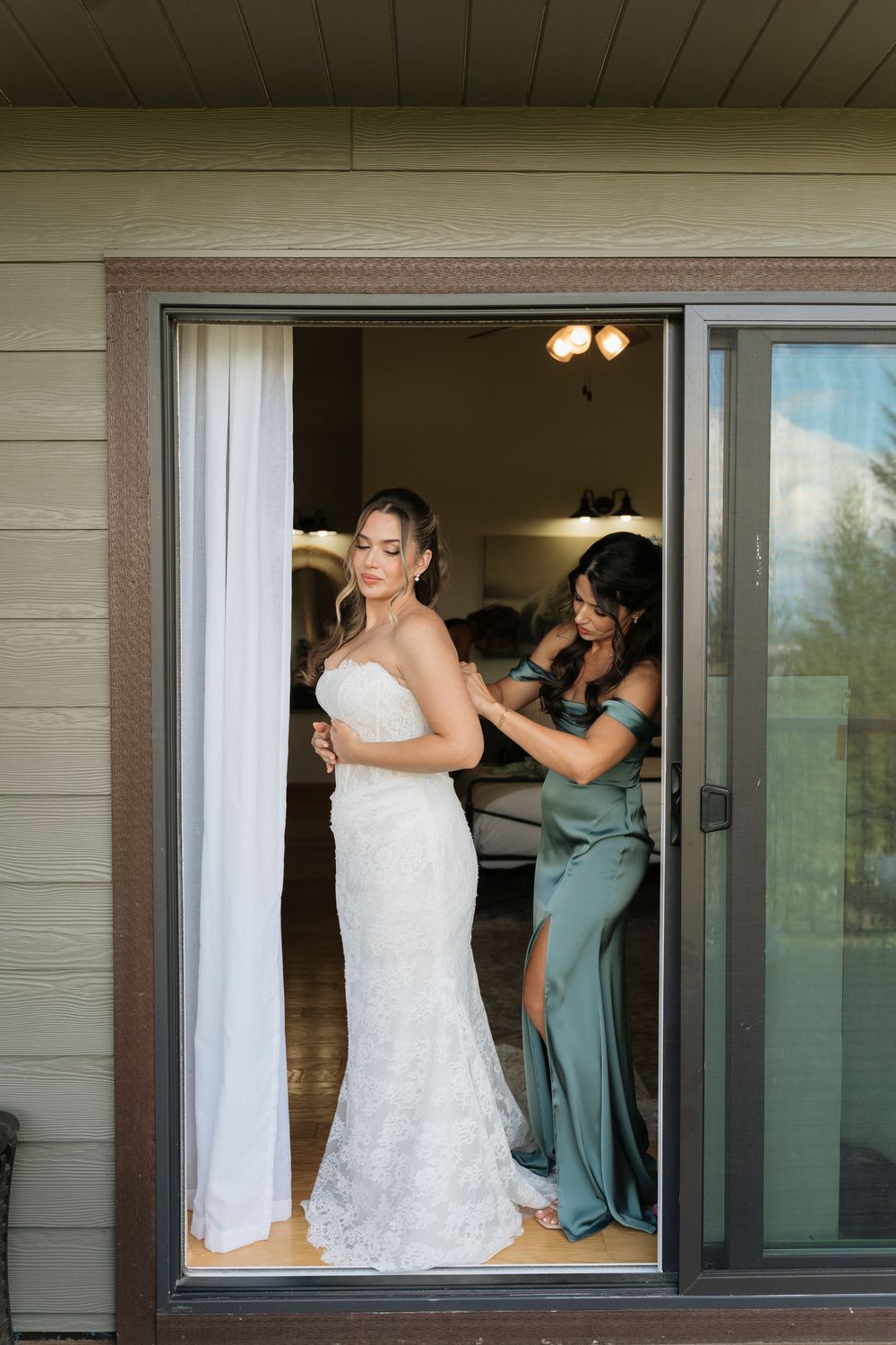 Bride in lace wedding gown being helped by bridesmaid in sage green dress at a glass door before the ceremony.