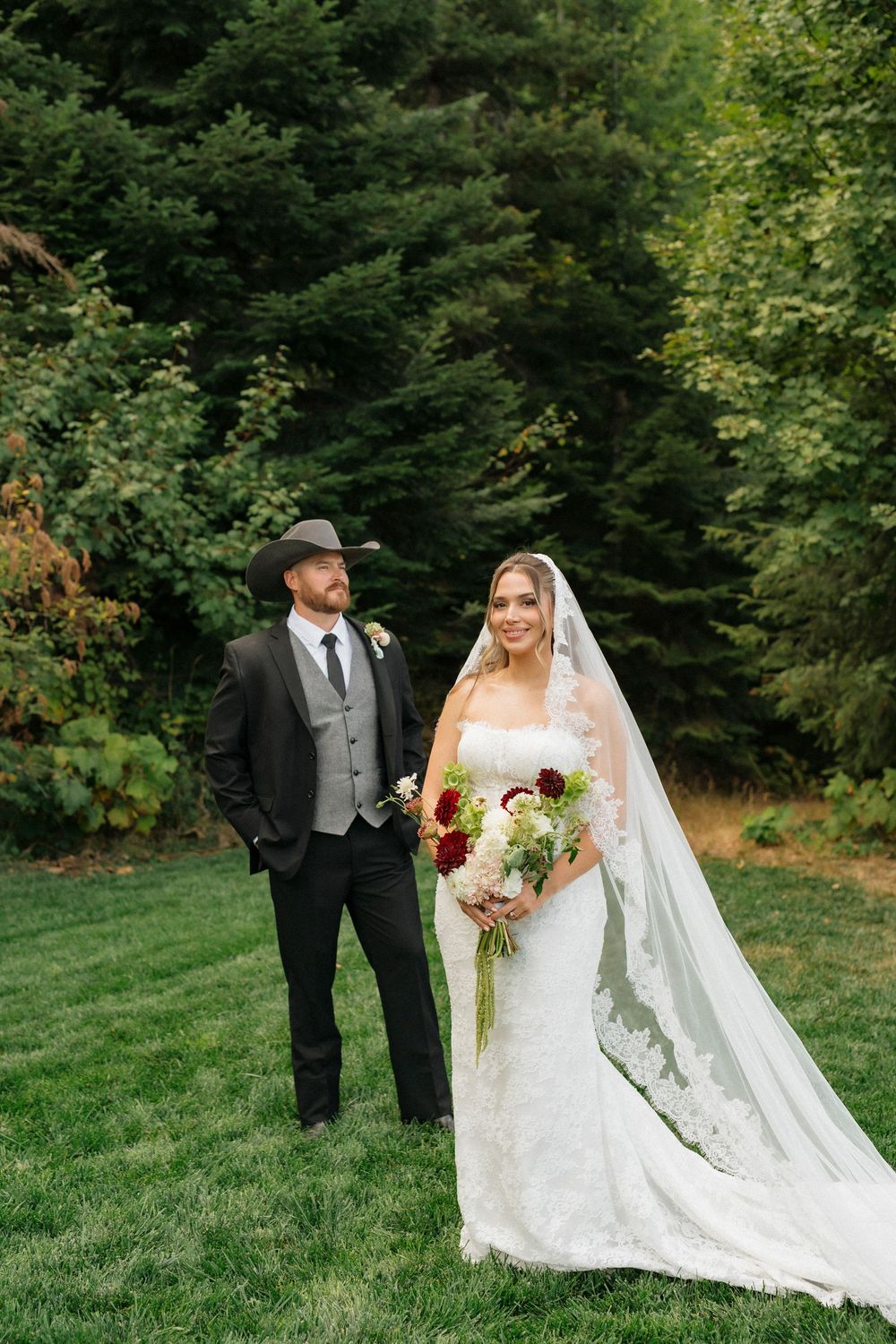 Bride in lace gown with veil holds bouquet beside groom in black suit and cowboy hat on green lawn.
