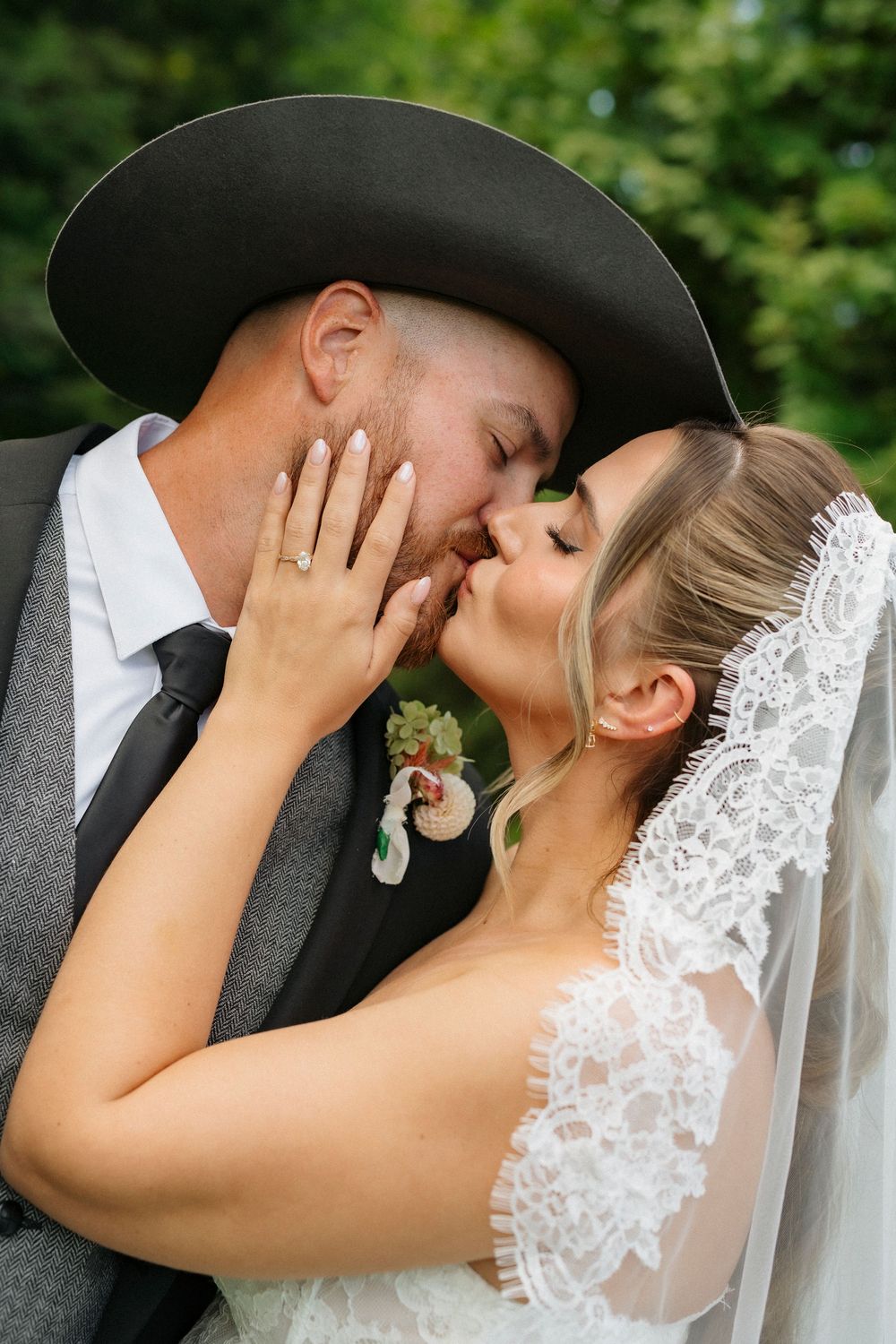 Bride and groom sharing a romantic kiss, groom wearing a black cowboy hat and gray suit with boutonniere.