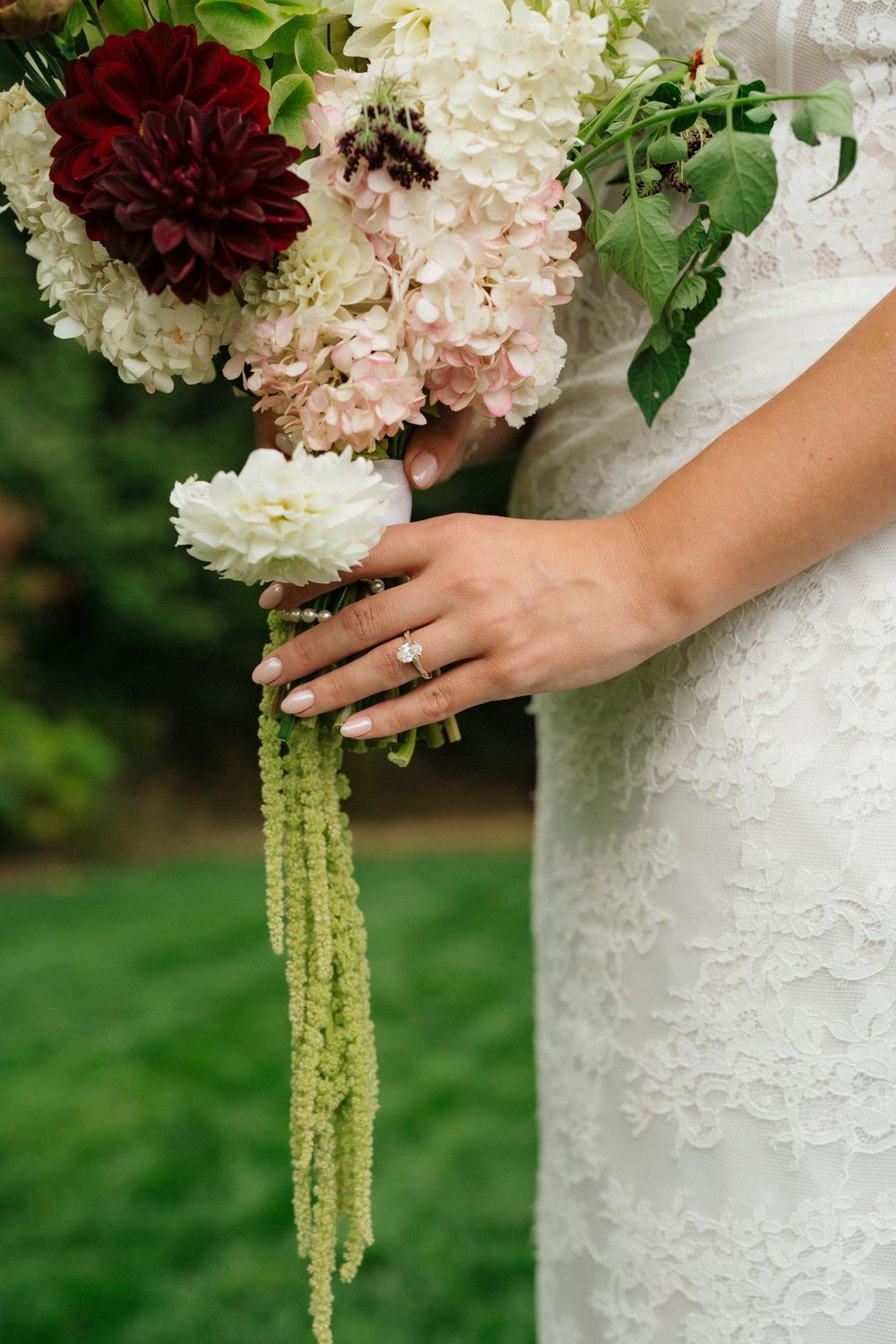 Bride in lace wedding dress holding bouquet of white, blush, and burgundy flowers with green amaranthus trails.