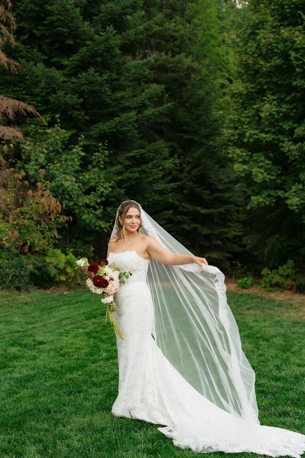Bride in lace mermaid gown holding bouquet and flowing veil outdoors on green lawn with forest backdrop.