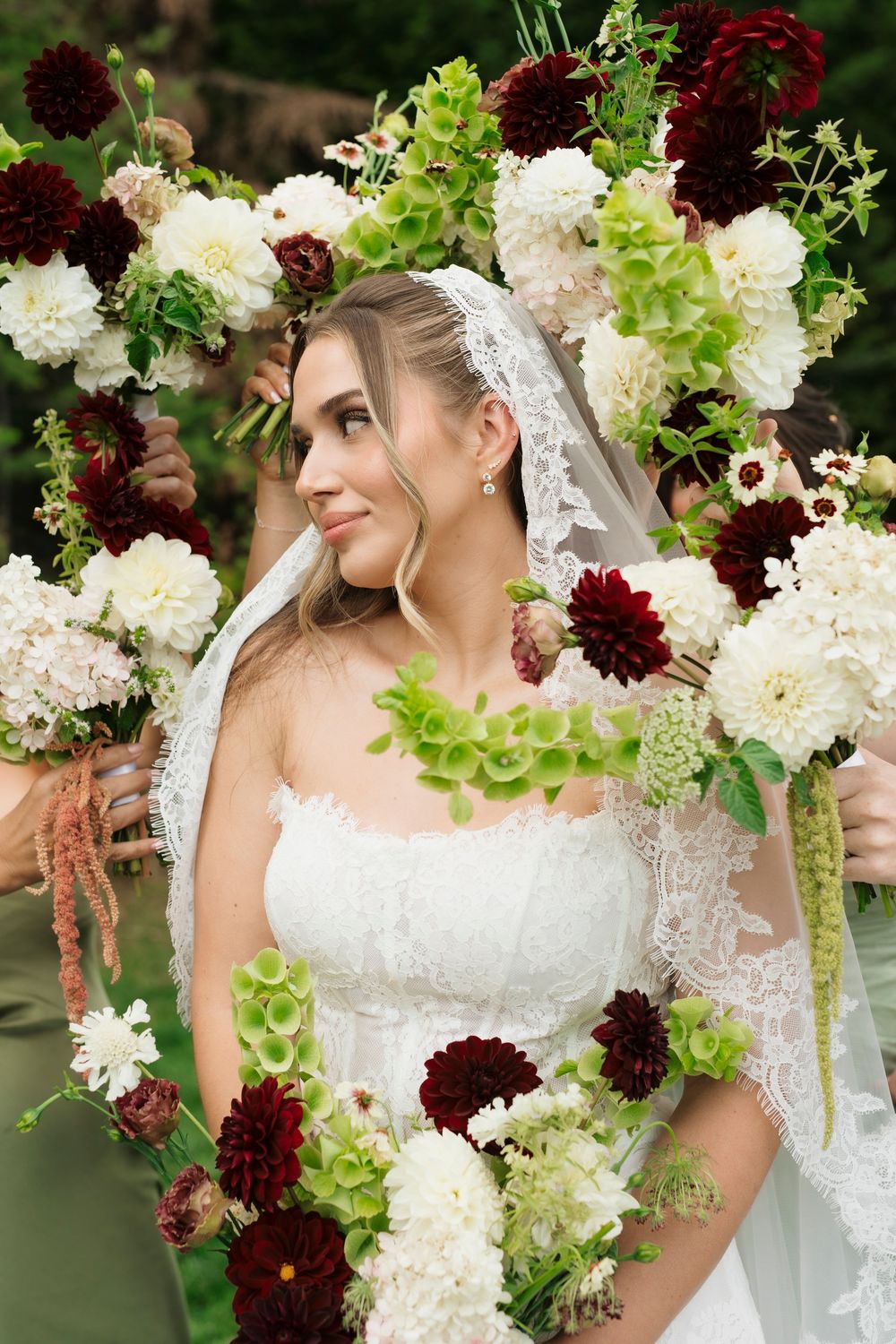 Bride in white lace gown and veil surrounded by lush floral arch with burgundy dahlias and white blooms.