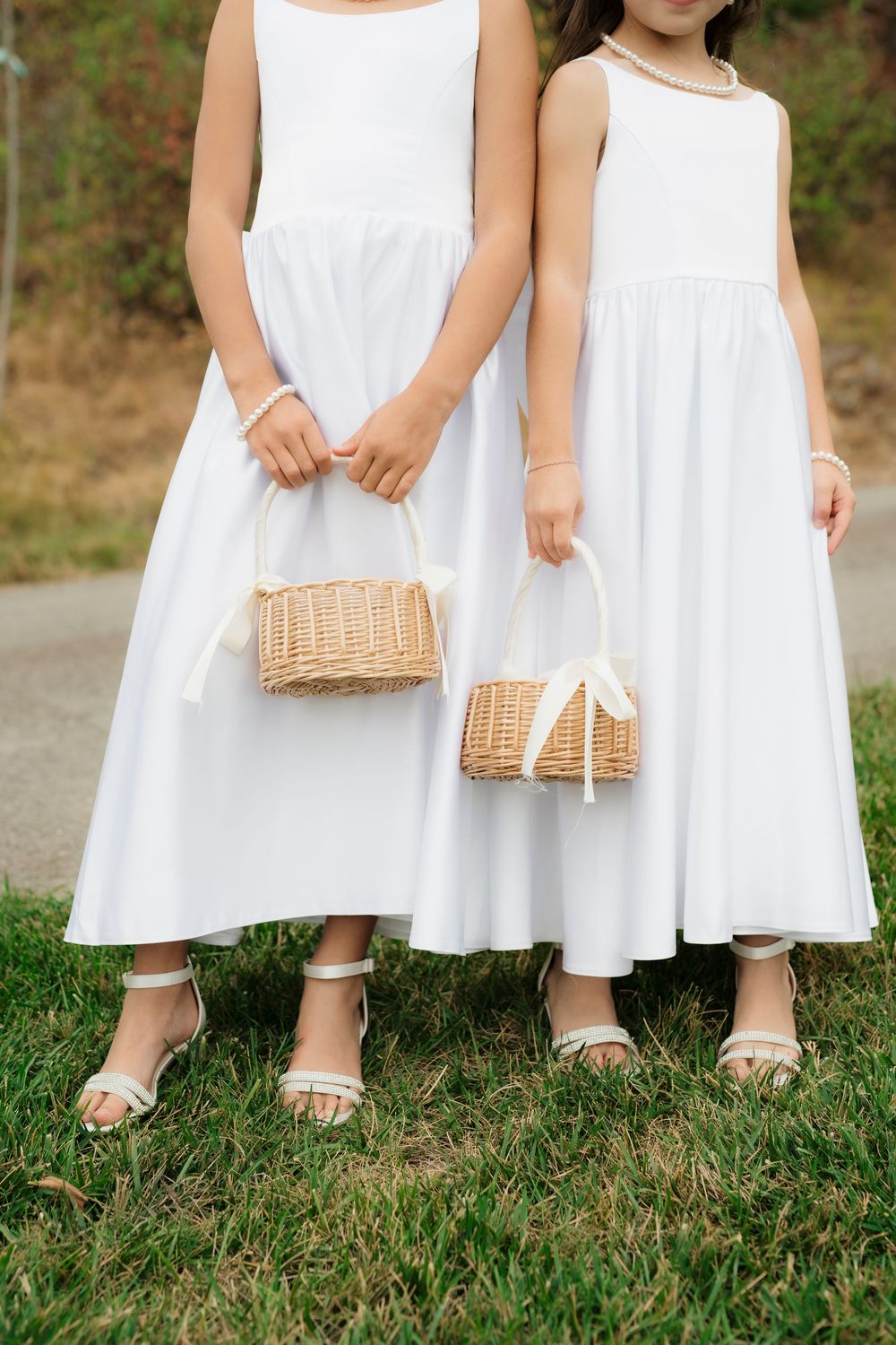 Two flower girls in white dresses holding wicker baskets with ribbon bows at an outdoor wedding.