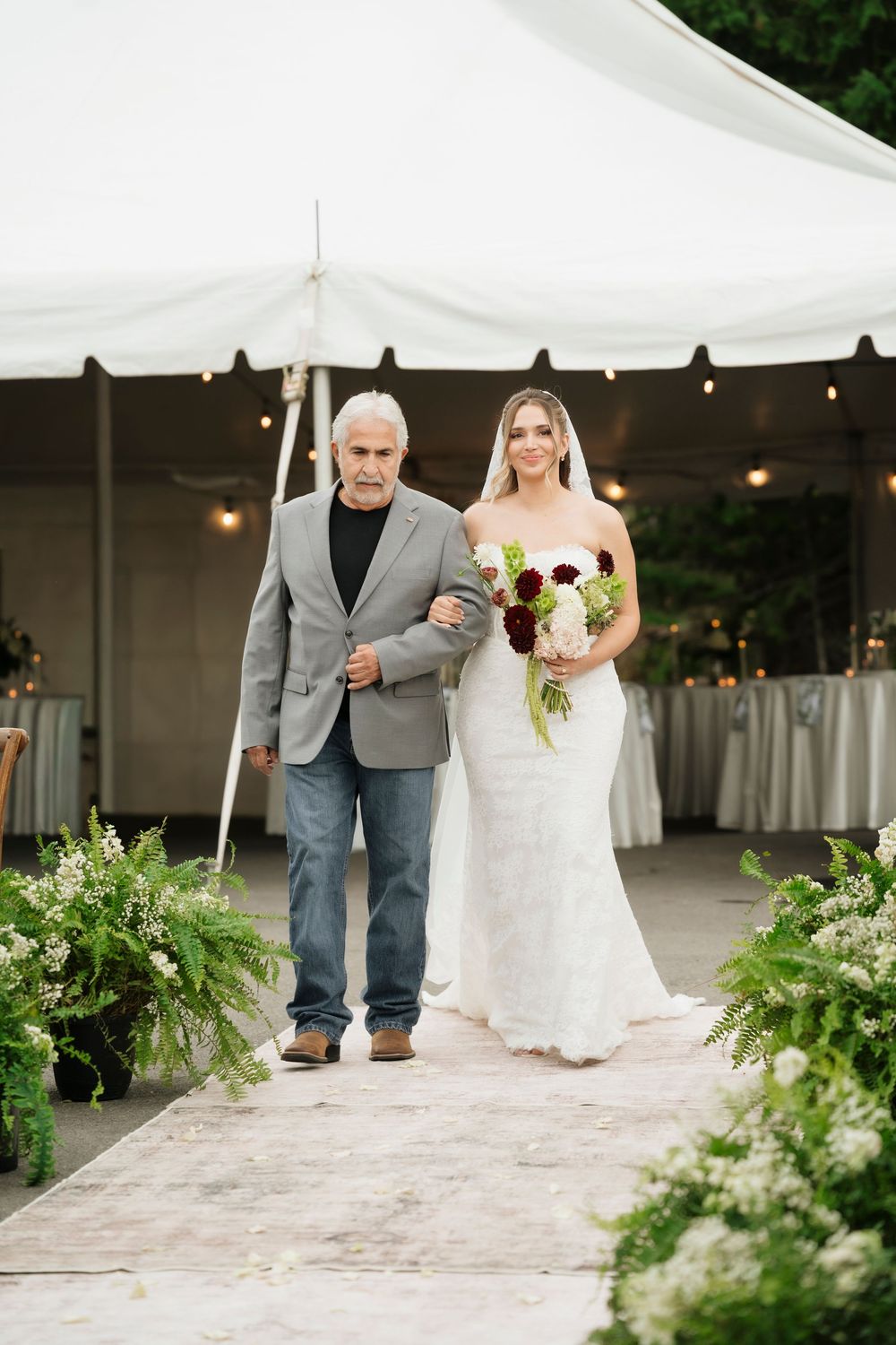 A bride in a white lace gown holds a bouquet while walking down the aisle with an older man at an outdoor wedding.