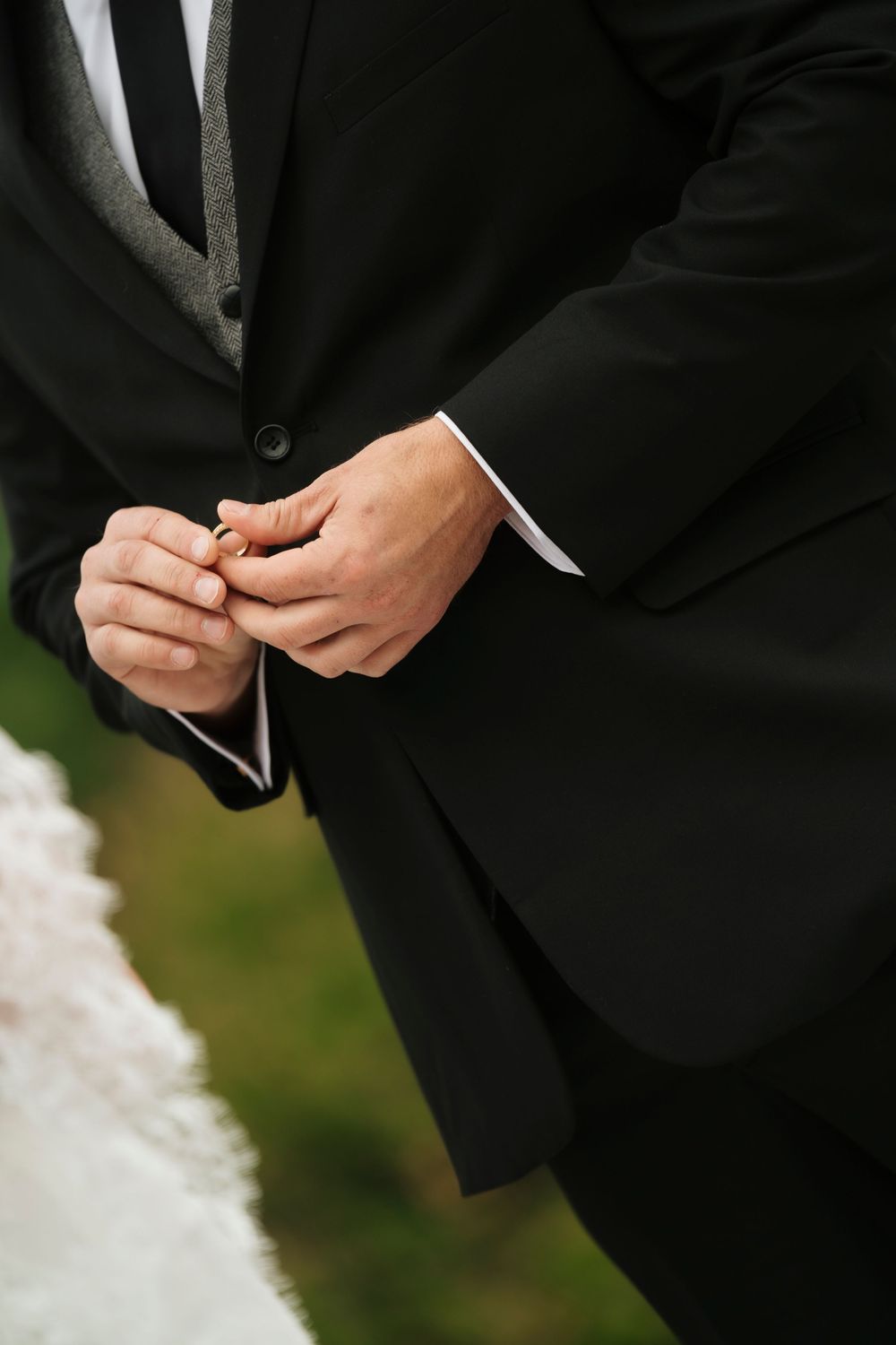 Groom in black suit holding a gold wedding ring during an outdoor wedding ceremony.