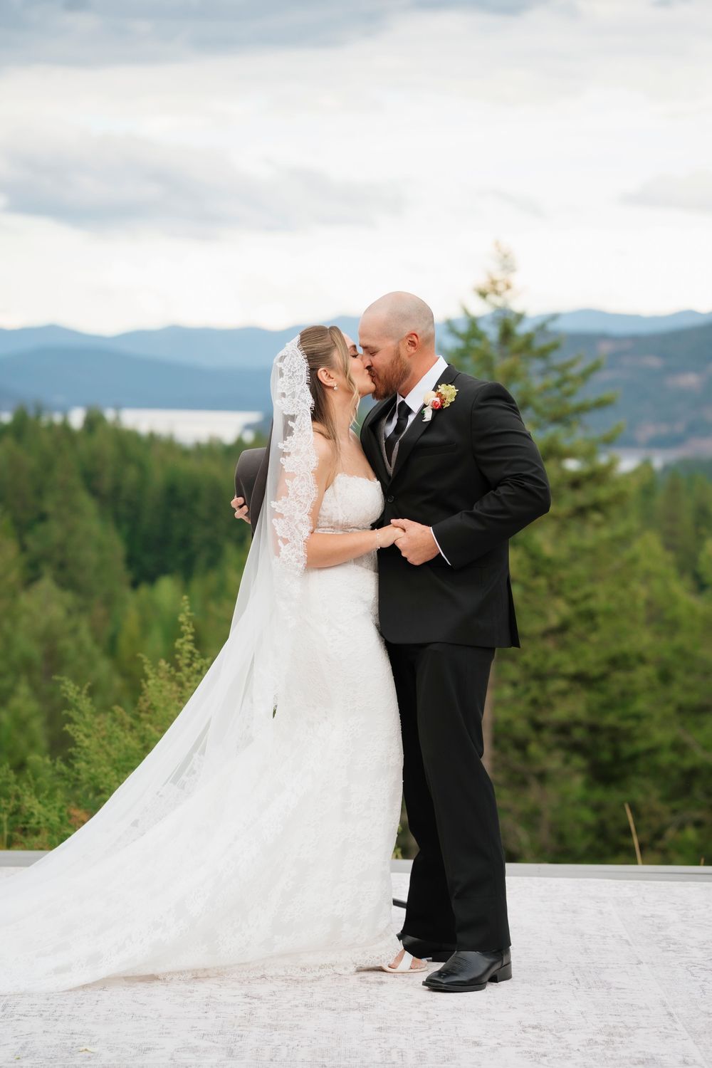 Bride and groom share a romantic kiss outdoors with mountain and lake views behind them on their wedding day.