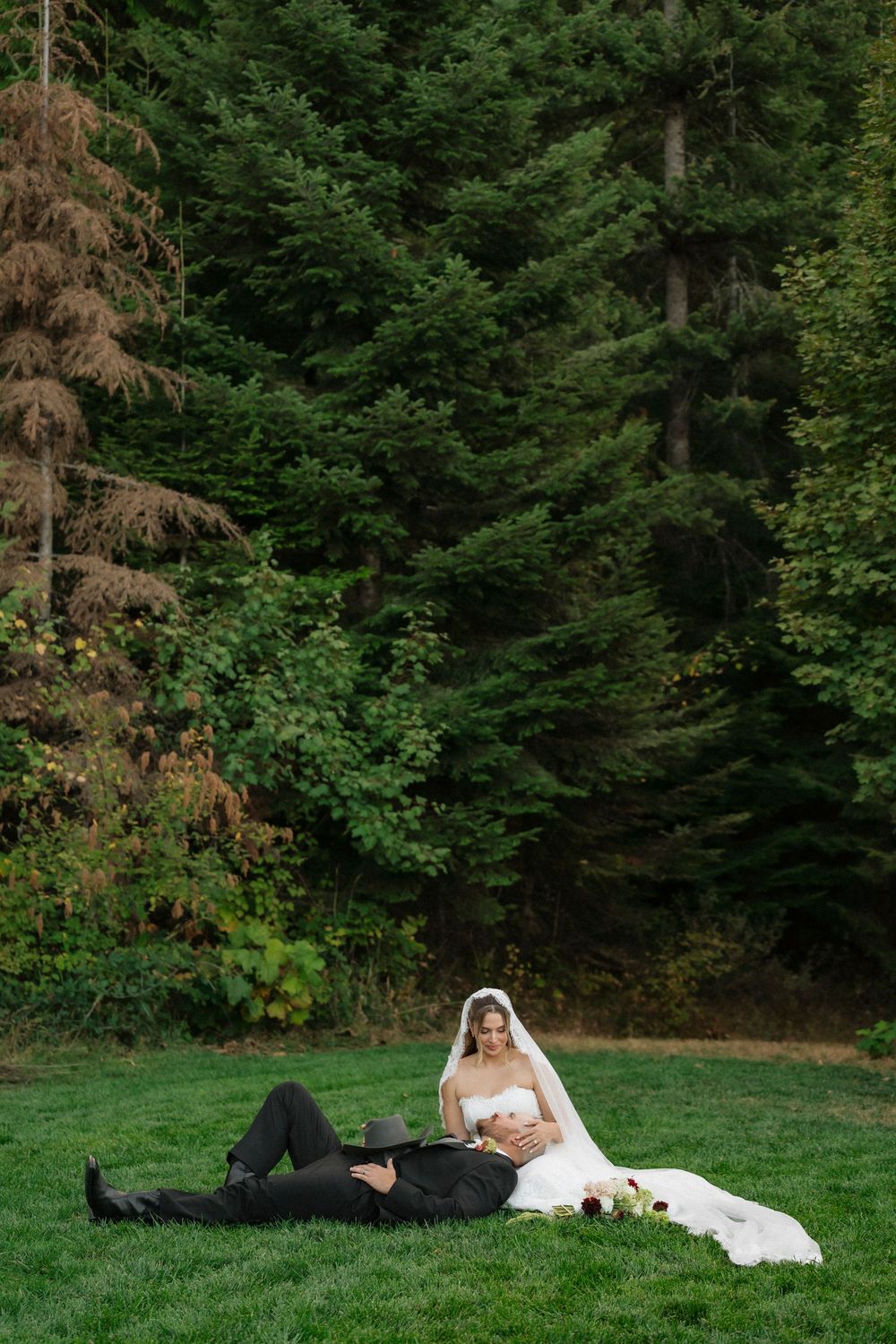 Bride and groom relaxing on green grass with lush forest backdrop during outdoor wedding photo session.