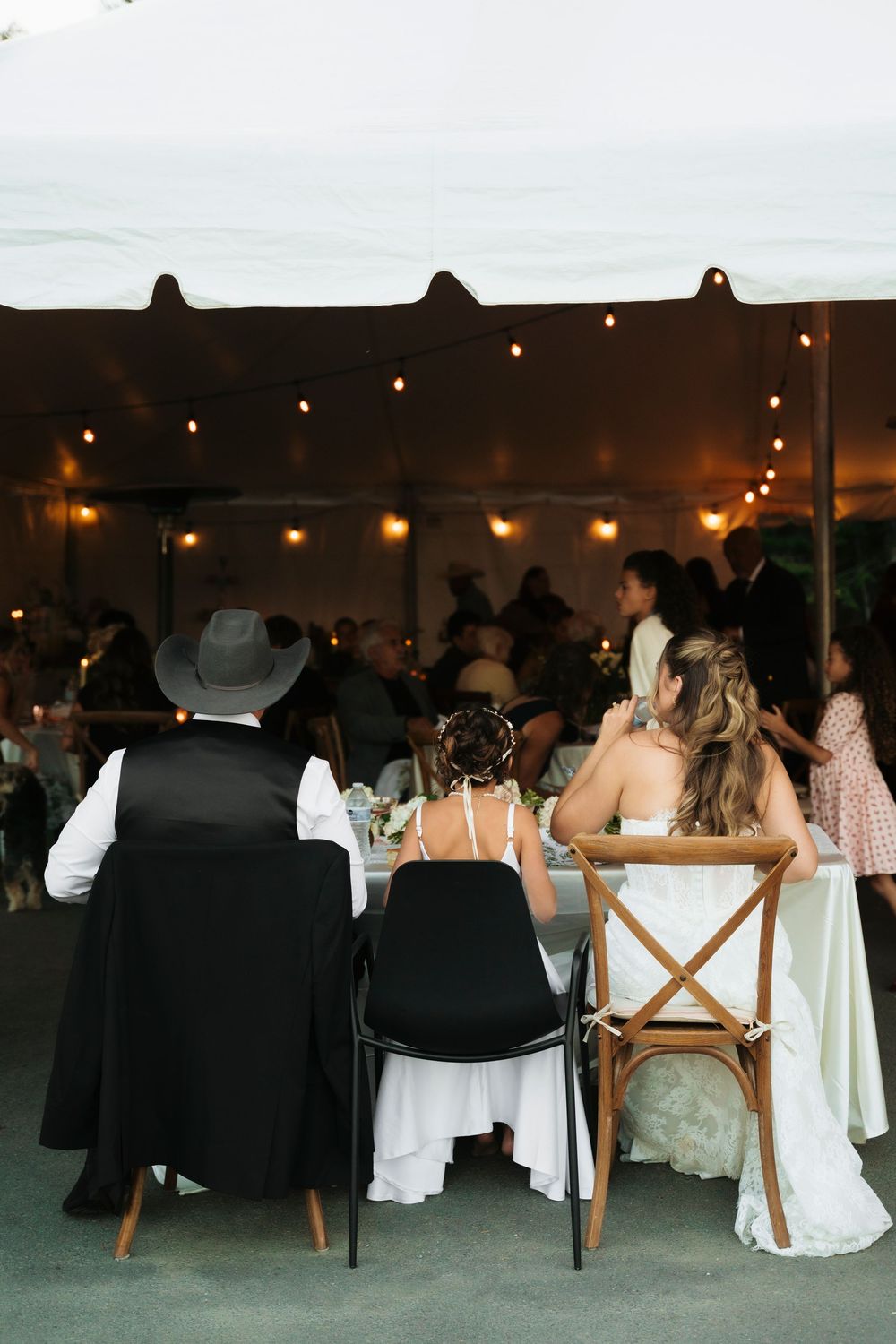 Wedding reception under a white tent with string lights, guests seated at tables, bride in white gown visible from behind.