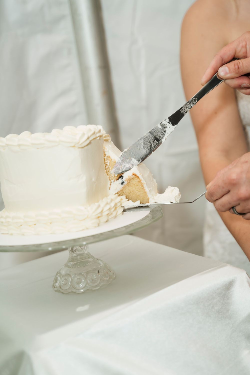 Bride and groom cutting a white wedding cake on a crystal cake stand with a silver serving knife.