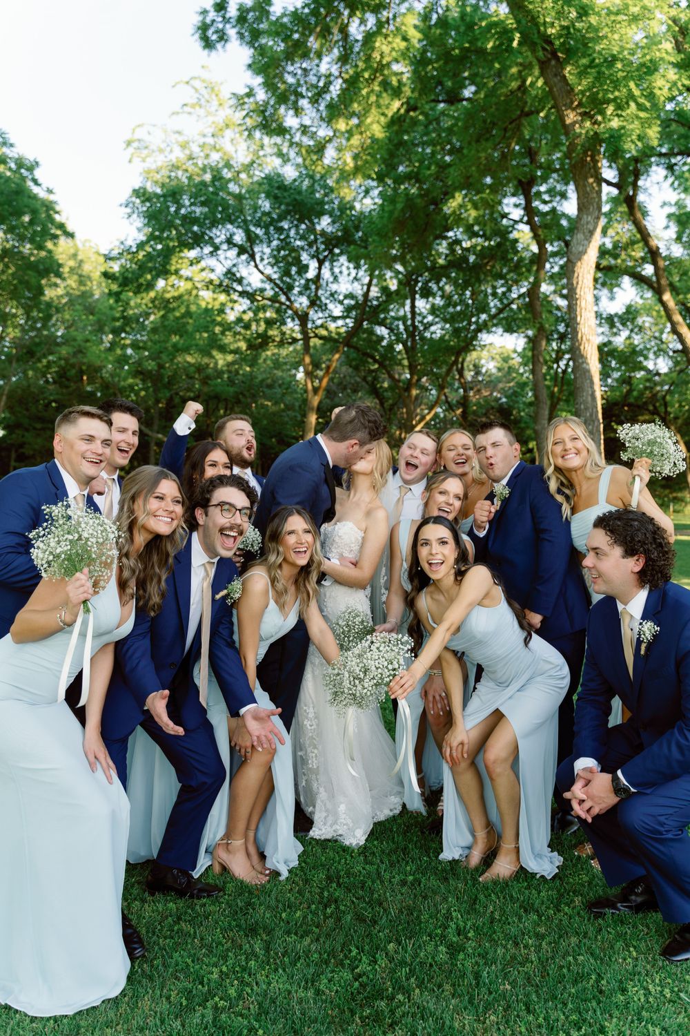 Wedding party poses playfully on grass in blue and gray attire.
