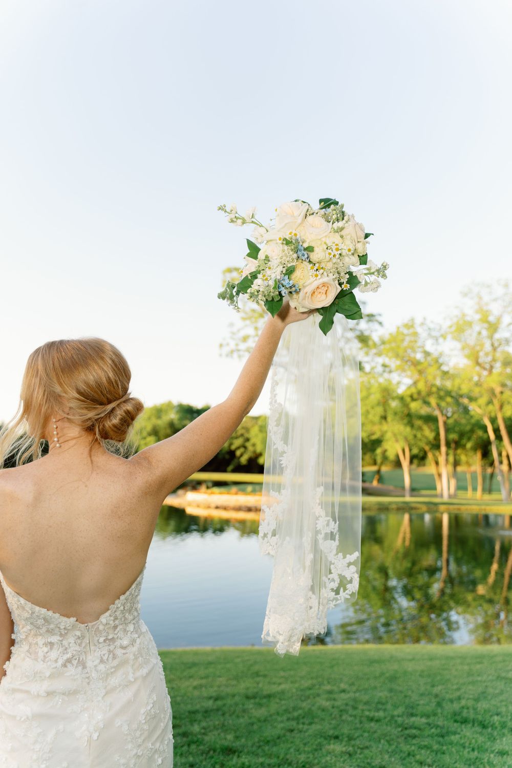 Bride in lace dress holding bouquet against sunset by a lake.
