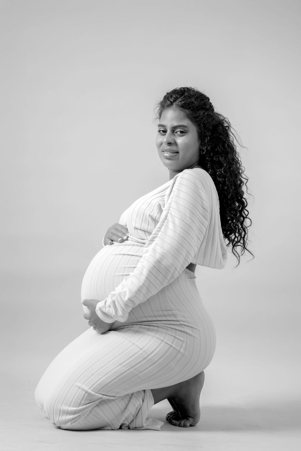 Maternity portrait of a woman in white dress kneeling against neutral background.