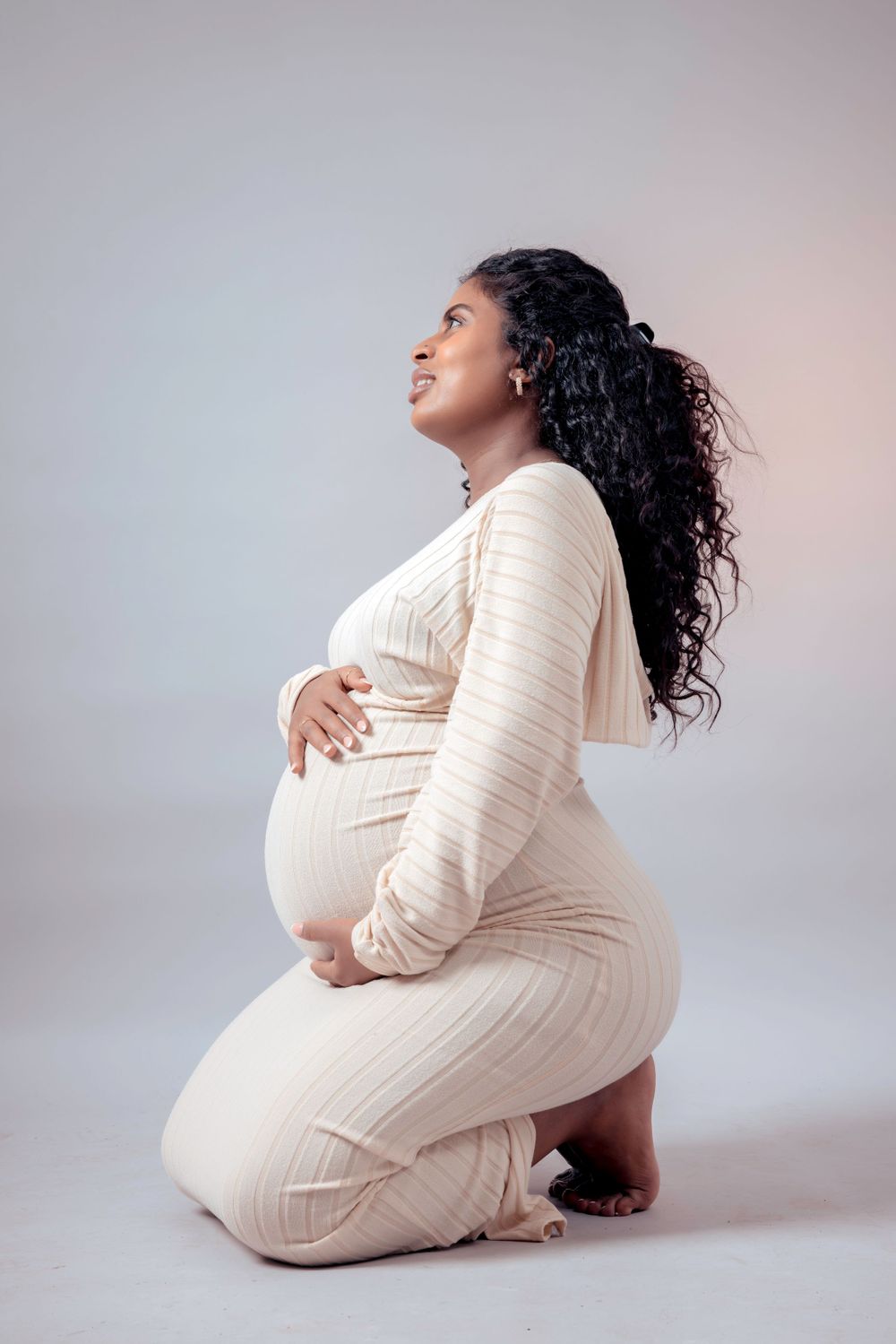 Side profile maternity photo of woman in fitted white dress against grey backdrop.