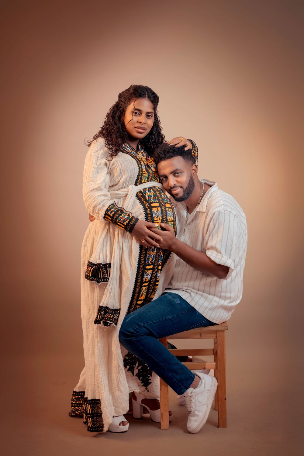 Studio maternity photo with couple in traditional dress against warm backdrop.