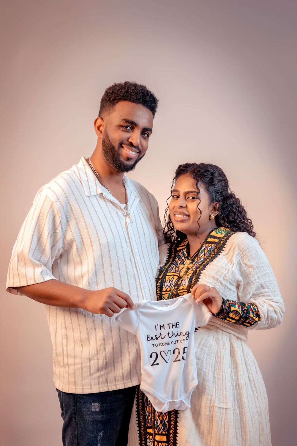 Expectant couple holding baby onesie while smiling in studio setting.