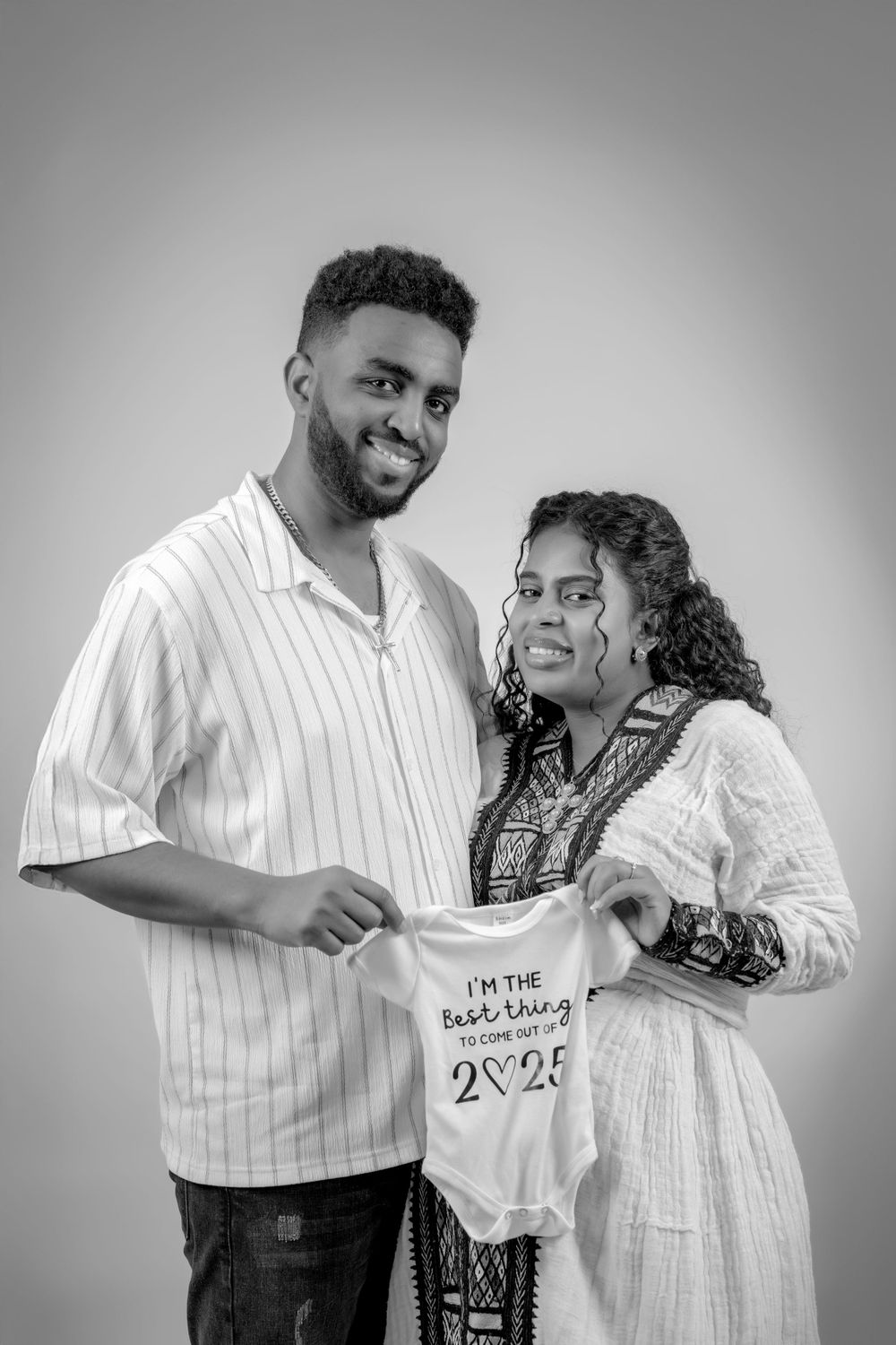 Couple holding baby onesie in black and white studio portrait.