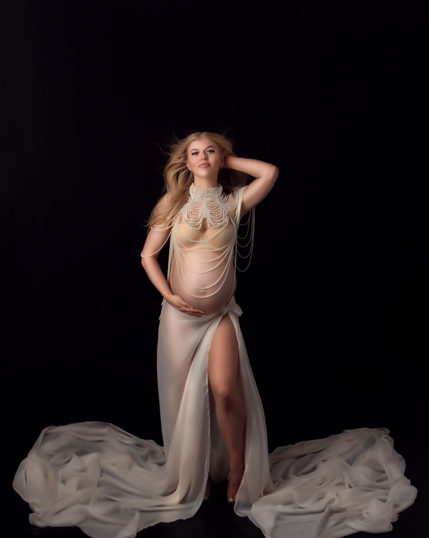 A pregnant mother in a white beaded bra necklace and silk fabric flowing bottom poses gracefully against a dark background.
