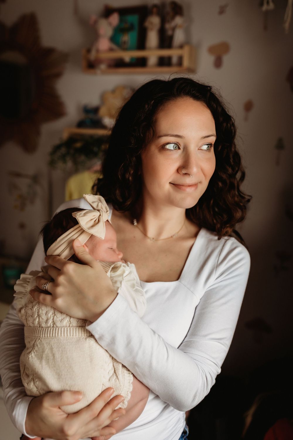 A woman in a white long-sleeve shirt holds a small baby wearing a cream-colored outfit with a bow headband.