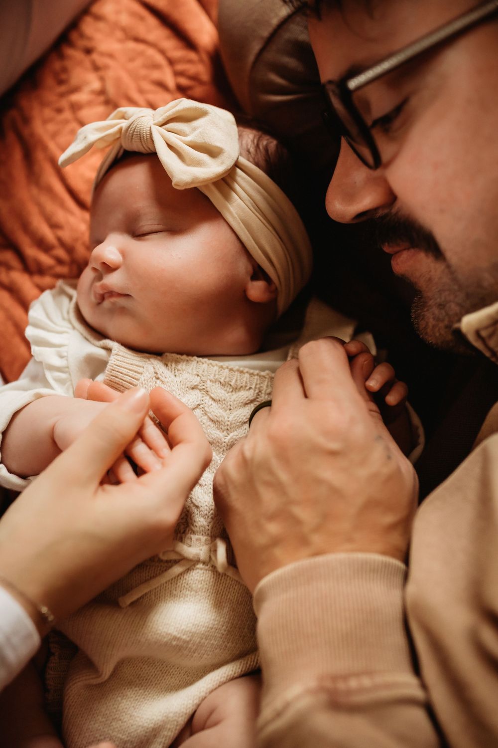 A sleeping newborn wearing a cream bow headband is held tenderly while wearing a knit outfit.