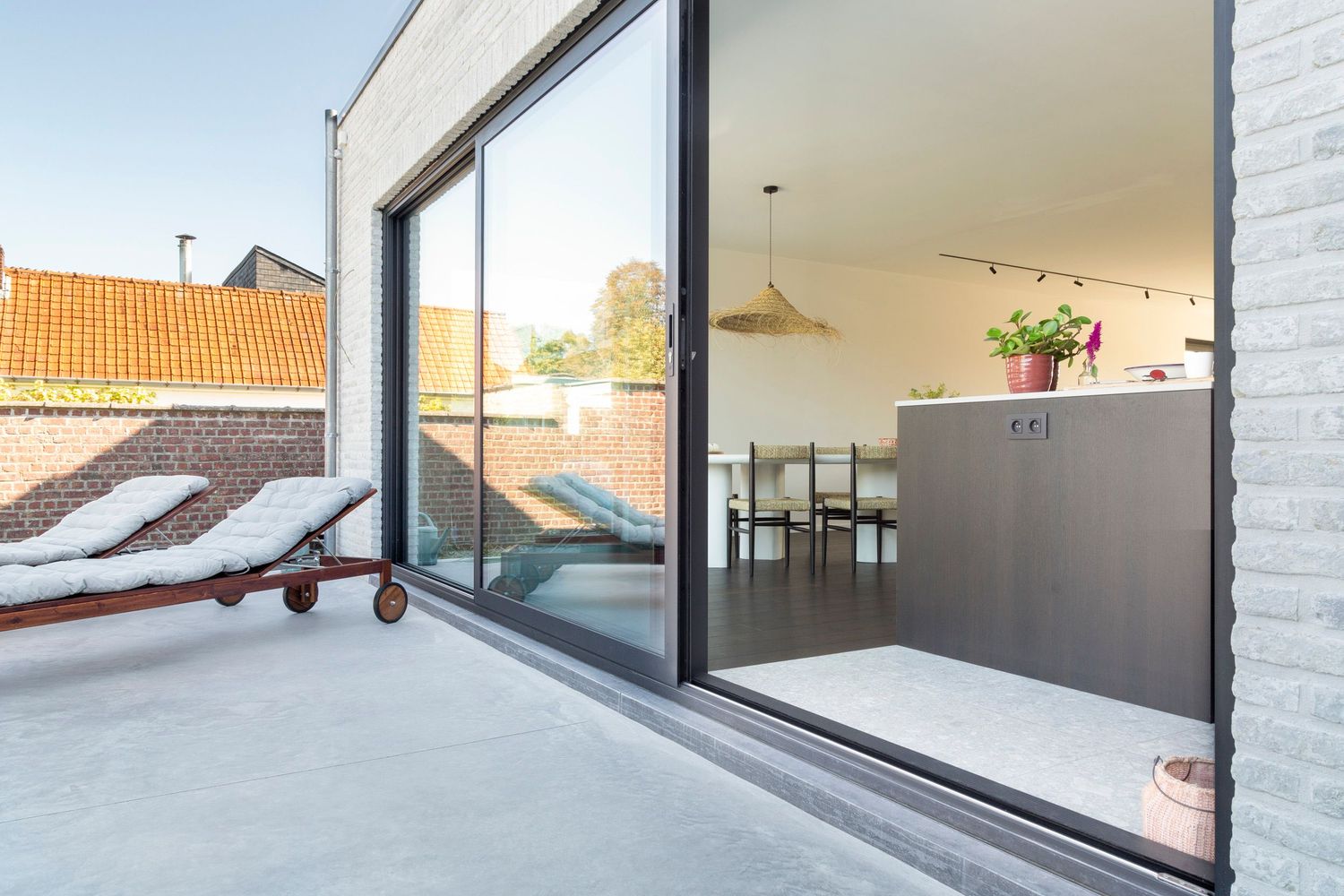 Outdoor patio with wooden lounge chair, white cushions, and floral arrangement against white brick wall.