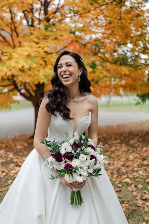 A joyful bride in a white gown holds a burgundy and white floral bouquet against vibrant autumn foliage.