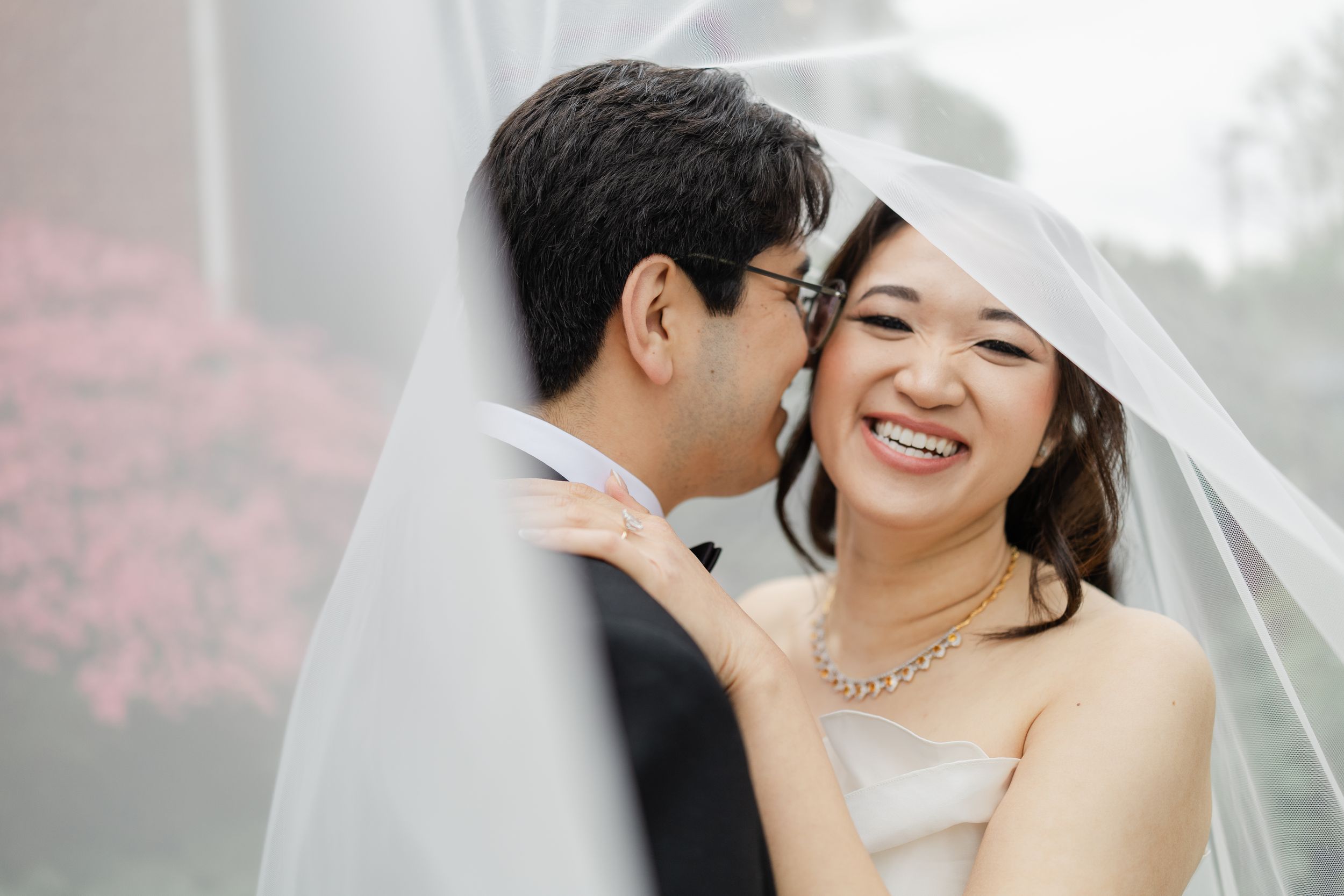 Groom kissing bride on cheek under wedding veil, bride smiling joyfully, pink flowers in background.