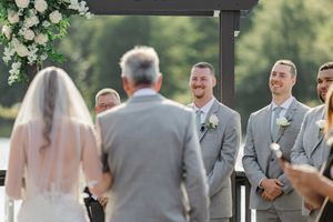 Wedding party in gray suits and light colored dresses stand in line during an outdoor ceremony.