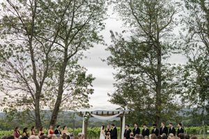 An outdoor wedding ceremony takes place under a white arbor surrounded by tall trees with guests seated on both sides.