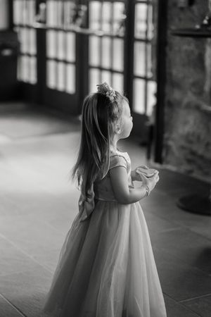 A small child in a flowing white dress gazes up at tall windows in a black and white photograph.
