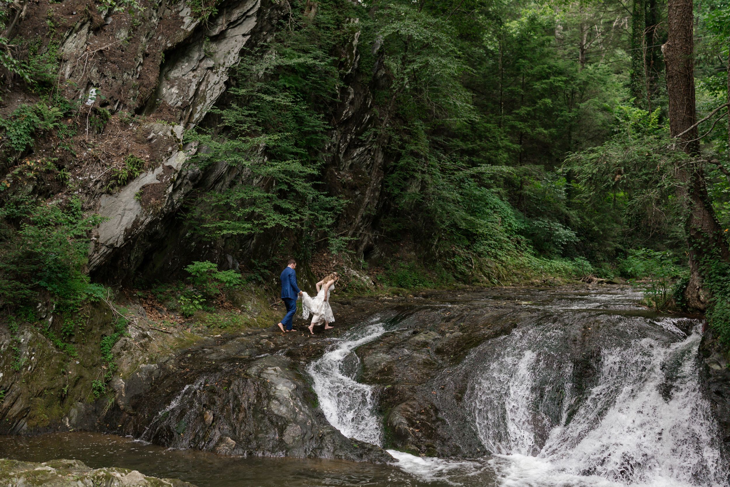 Person and dog exploring rocky waterfall surrounded by lush green forest trees.