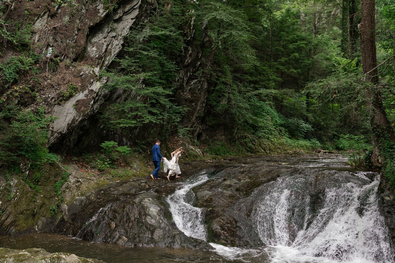 Person and dog exploring rocky waterfall surrounded by lush green forest trees.