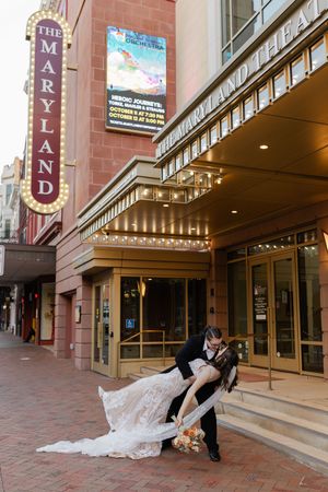 A romantic couple shares a dip kiss pose outside a theater entrance with illuminated marquee lights.