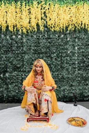 Traditional bridal scene with yellow dupatta against green wall backdrop and gold decorative elements on white cloth setting.