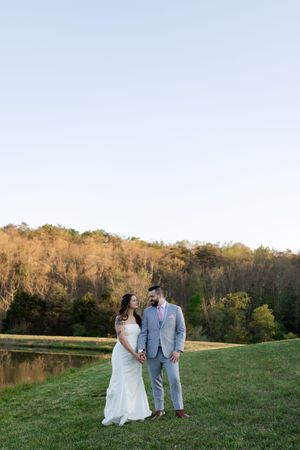 A bride and groom hold hands and smile at each other on a green lawn with a pond and autumn trees at golden hour.