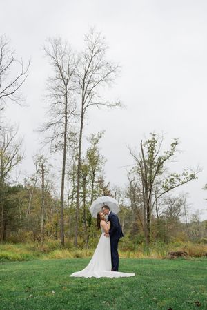 A couple embraces on a hilltop meadow surrounded by bare trees on an overcast autumn day.