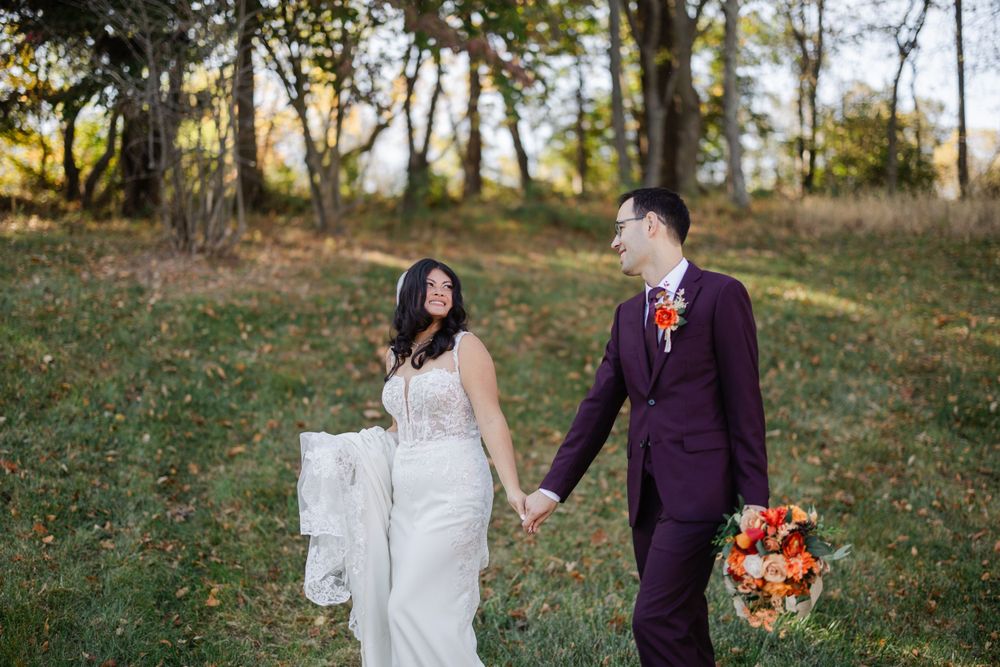 A couple in wedding attire walks hand in hand through an autumn forest setting, with the groom carrying a vibrant bouquet.