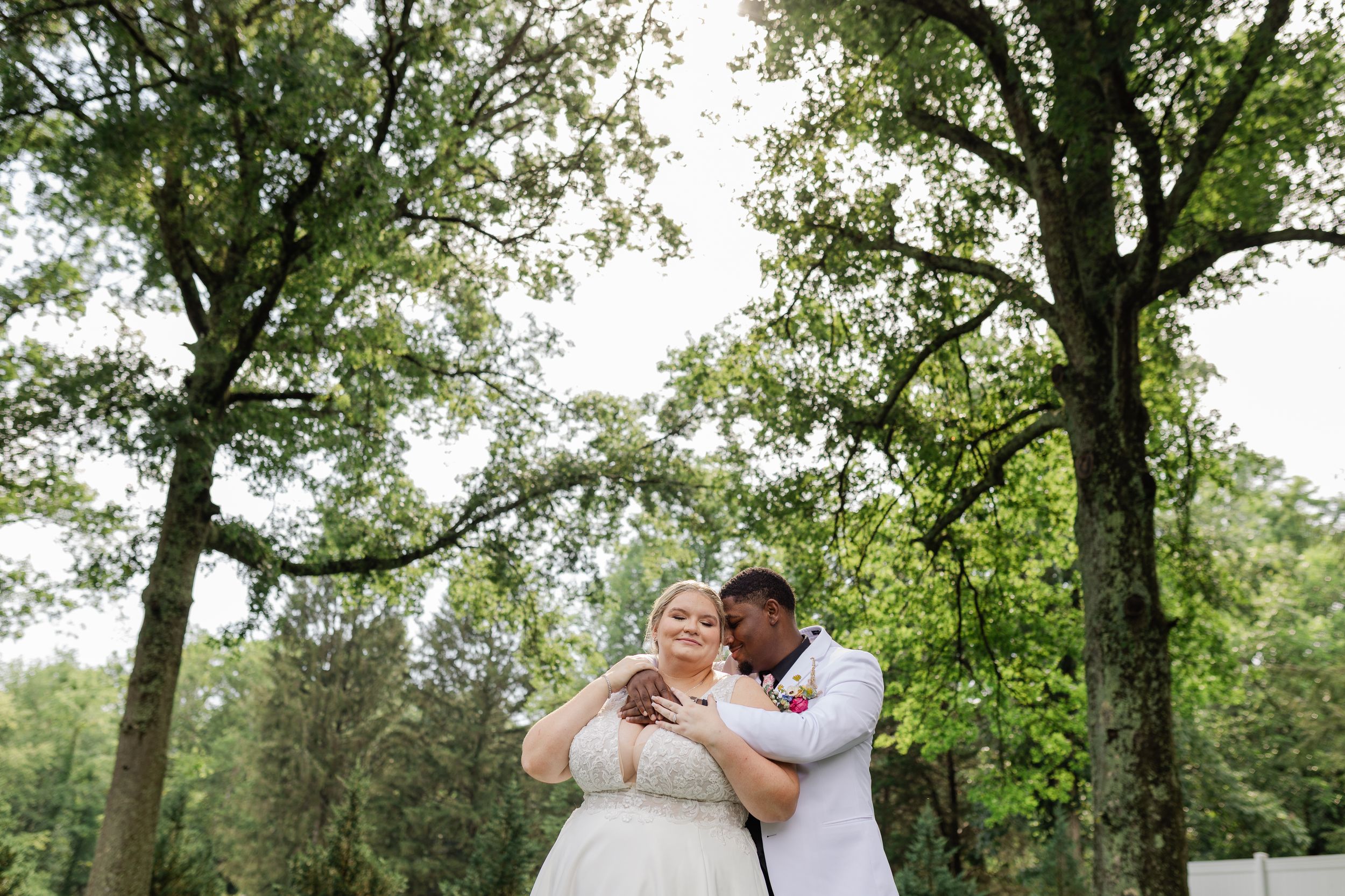 A romantic wedding couple shares a tender moment under tall green trees during their outdoor celebration.