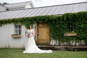 Bride in white lace gown holding colorful bouquet poses before ivy-covered white barn with wooden door.