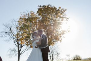 Couple in wedding attire embraces under autumn trees during sunset while photographers capture their special moment.