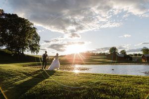 Bride and groom walk hand-in-hand by a sunlit pond at golden hour on a lush green golf course.