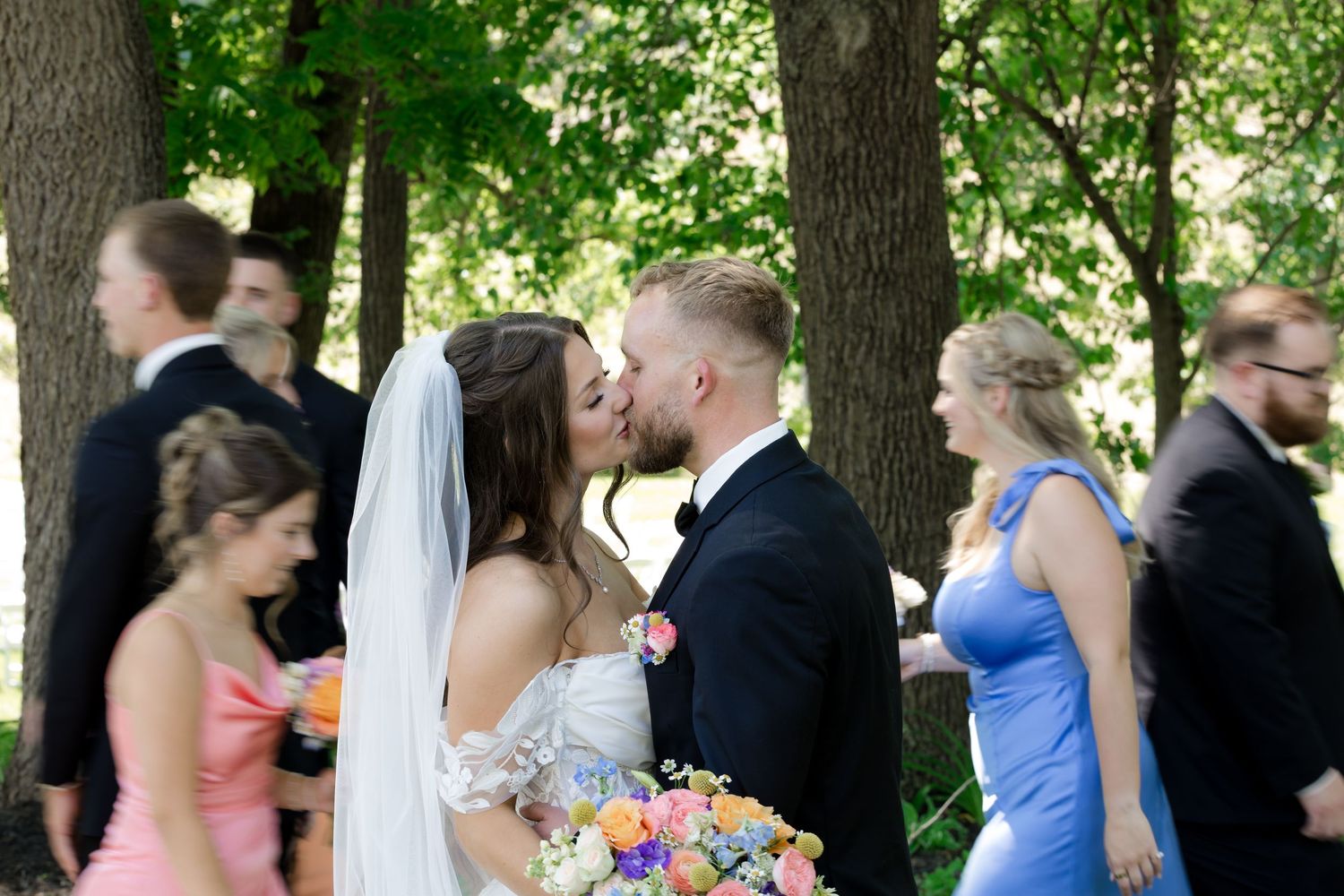 Bride and groom share a kiss outdoors surrounded by wedding party in colorful attire under lush green trees.